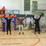 Trzech dorosłych mężczyzn odgrywa scenę na sali gimnastycznej – w tle zamek z wieżą, z której wygląda księżniczka, oraz dekoracje lasu i drzewa z papierowymi pszczołami.