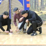 Trzech mężczyzn w strojach scenicznych, z opaskami z czułkami, kuca na scenie i skupia się na manipulowaniu małymi przedmiotami w pudełkach, odgrywając scenę z przedstawienia.