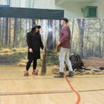 Na tle scenografii przedstawiającej las stoi kobieta przebrana za czarownicę z miotłą, w czarnym stroju i kapeluszu, oraz mężczyzna w stroju Szewczyka Dratewki; obok leży dekoracja przypominająca górę z oczami.