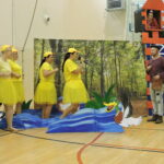 Cztery kobiety przebrane za kaczuszki w żółtych strojach i czapkach z dziobami występują na scenie przed dekoracją rzeki i lasu; jedna z nich śpiewa do mikrofonu. Obok stoi mężczyzna jako Szewczyk Dratewka, a z wieżyczki dekoracyjnego zamku spogląda postać księżniczki.