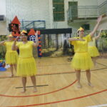 Cztery kobiety przebrane za kaczuszki w żółtych strojach i czapkach z dziobami występują na scenie przed dekoracją rzeki i lasu; jedna z nich śpiewa do mikrofonu. Obok stoi mężczyzna jako Szewczyk Dratewka, a z wieżyczki dekoracyjnego zamku spogląda postać księżniczki.