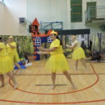 Cztery kobiety przebrane za kaczuszki w żółtych strojach i czapkach z dziobami występują na scenie przed dekoracją rzeki i lasu; z wieżyczki dekoracyjnego zamku spogląda postać księżniczki.