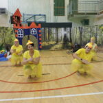 Cztery kobiety przebrane za kaczuszki w żółtych strojach i czapkach z dziobami występują na scenie przed dekoracją rzeki i lasu; z wieżyczki dekoracyjnego zamku spogląda postać księżniczki.