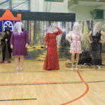 cztery kobiety w kolorowych sukniach i z woalkami na twarzach stoi na scenie szkolnej w dekoracji lasu; w tle czarownica z miotłą oraz bajkowy zamek