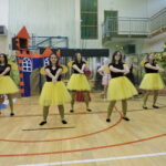 Pięć kobiet przebranych za pszczoły tańczy na tle dekoracji leśnej i zamku; ubrane są w żółte spódniczki, czarne koszulki i opaski z czułkami.