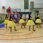 Pięć kobiet przebranych za pszczoły tańczy na tle dekoracji leśnej i zamku; ubrane są w żółte spódniczki, czarne koszulki i opaski z czułkami.