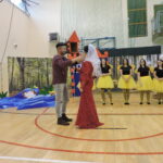 Mężczyzna w czapce zakłada welon kobiecie w czerwonej sukni podczas sceny z przedstawienia; w tle kobiety przebrane za pszczoły oraz dekoracje lasu i zamku.