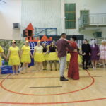 Para w centrum sali gimnastycznej tańczy, wokół nich uczestnicy przedstawienia w barwnych kostiumach, trzymają się za ręce tworząc krąg.