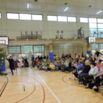 Uczestnicy przedstawienia tanecznym krokiem wbiegają na środek sali gimnastycznej, trzymając się za ręce; publiczność, w tym dzieci i dorośli, siedzi na krzesłach i nagradza ich brawami.