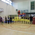 Grupa dorosłych występujących w kolorowych kostiumach stoi w szeregu na zakończenie przedstawienia w sali gimnastycznej; w tle dekoracja lasu i bajkowego zamku.