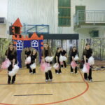 Grupa dziewczynek w czarnych strojach cheerleaderek z biało-różowymi pomponami wykonuje układ taneczny na sali gimnastycznej przed dekoracją leśną i bajkowym zamkiem.