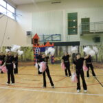 Grupa dziewczynek w czarnych strojach cheerleaderek z biało-różowymi pomponami wykonuje układ taneczny na sali gimnastycznej przed dekoracją leśną i bajkowym zamkiem.