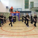 Dziewczynki w czarnych strojach cheerleaderek z ozdobnymi paskami wykonują układ taneczny na tle dekoracji przedstawiającej las i bajkowy zamek na sali gimnastycznej.