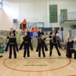 Dziewczynki w czarnych strojach cheerleaderek z ozdobnymi paskami wykonują układ taneczny na tle dekoracji przedstawiającej las i bajkowy zamek na sali gimnastycznej.