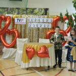 Dwoje dzieci w eleganckich strojach prowadzi „Koncert Życzeń” na tle złotej kurtyny i czerwonych balonów w kształcie serc, obok dekoracji z logo „TVP Lubomierz”.