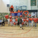 Grupa dzieci występuje na scenie z czerwonymi balonami w kształcie serc; w tle dekoracja zamku, a z boku dzieci grają na akordeonie i skrzypcach.