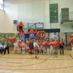 Grupa dzieci występuje na scenie z czerwonymi balonami w kształcie serc; w tle dekoracja zamku, a z boku dzieci grają na akordeonie i skrzypcach.