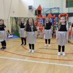 Grupa dziewczynek ubranych w czarne koszulki i srebrne spódnice występuje tanecznie na sali gimnastycznej; w tle dzieci trzymają czerwone balony w kształcie serc.