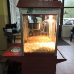 Maszyna do popcornu z logo Banku Spółdzielczego w Mszanie Dolnej, wypełniona świeżo uprażonym popcornem, ustawiona wewnątrz budynku szkolnego.