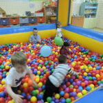 Dzieci bawią się radośnie w kolorowym basenie z kulkami, wypełnionym setkami wielobarwnych piłeczek. Wśród nich unoszą się balony, a całe miejsce znajduje się w przestrzeni sali przedszkolnej zabezpieczonej siatką.