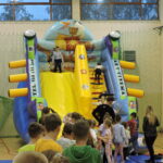 Dzieci wspinają się i zjeżdżają z dużej, kolorowej dmuchanej zjeżdżalni w kształcie statku pirackiego, ustawionej w sali gimnastycznej. Przed zjeżdżalnią ustawiła się kolejka dzieci, a dorośli czuwają nad bezpieczeństwem zabawy.