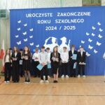 Grupa uczniów i uczennic w odświętnych strojach stoi na tle niebieskiej dekoracji z napisem „Uroczyste zakończenie roku szkolnego 2024/2025”; po prawej nauczycielka przemawia przy mównicy.