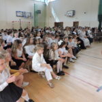 Publiczność w sali gimnastycznej, dzieci i dorośli siedzą w rzędach, patrząc w stronę sceny; najmłodsi uczniowie klas I–III siedzą z przodu i biją brawo.