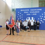 Poczet sztandarowy z uczniami w galowych strojach stoi na sali gimnastycznej przed dekoracją zakończenia roku; za nimi grupa młodzieży z klas starszych.