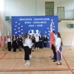 Przekazanie sztandaru przez uczniów; dwie grupy uczniów stoją naprzeciw siebie na tle dekoracji z napisem „Zakończenie roku szkolnego 2024/2025”.