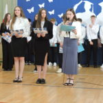 Trzy uczennice w galowych strojach stoją na pierwszym planie; jedna z nich czyta z kartki do mikrofonu, pozostałe trzymają dyplomy z napisem „Pożegnania”.