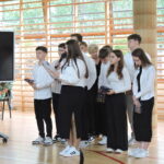 Grupa uczniów w białych koszulach i ciemnych spodniach stoi przy oknach sali gimnastycznej; jedna z uczennic przemawia do mikrofonu, inni trzymają upominki.