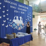 Dwie nauczycielki prowadzą uroczystość zakończenia roku szkolnego; stoją przy mikrofonach na tle niebieskiej dekoracji z sylwetkami absolwentów, kwiatami i motylami. Na pierwszym planie stół z ułożonymi nagrodami.