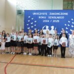 Grupa uczniów i uczennic z klas I–III stoi na tle dekoracji zakończenia roku; każde dziecko trzyma książkę w rękach, obok stoją wychowawczynie, rodzice i dyrekcja szkoły.