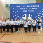 Uczniowie klasy IV pozują do zdjęcia z nagrodami książkowymi na tle niebieskiej dekoracji zakończenia roku; po bokach stoją nauczycielki.