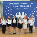 Sześcioro uczniów stoi w rzędzie z dyplomami za osiągnięcia, za nimi nauczycielki i dekoracja z napisem „Uroczyste zakończenie roku szkolnego 2024/2025”.