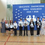 Grupa uczniów z nagrodami książkowymi pozuje do zdjęcia na tle dekoracji zakończenia roku szkolnego; po bokach stoją nauczycielki.