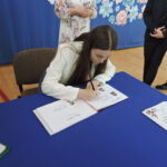 Uczennica siedzi przy stole i wpisuje się do Złotej Księgi, na stole widoczne strony z jej zdjęciem i nazwiskiem.