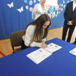 Uczennica siedzi przy stole i wpisuje się do Złotej Księgi, na stole widoczne strony z jej zdjęciem i nazwiskiem.