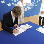 Uczeń w czarnym garniturze podpisuje się w Złotej Księdze; obok nauczycielka oraz inny uczeń oczekujący na swoją kolej.