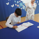 Uczeń podpisuje się w Złotej Księdze przy stole z niebieskim obrusem; nauczycielka z uśmiechem obserwuje moment wpisu.