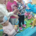 Dzieci w opaskach z symbolem kropli wody podają hipopotamowi niebieskie klocki jako jedzenie.