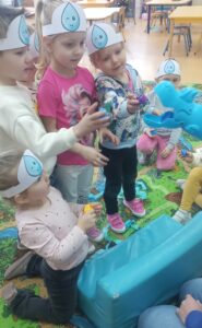 Dzieci w opaskach z symbolem kropli wody podają hipopotamowi niebieskie klocki jako jedzenie.