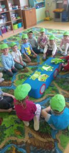 Dzieci w zielonych czapkach siedzą na dywanie i słuchają bajki o żabkch.