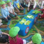 Dzieci w zielonych czapkach siedzą na dywanie i słuchają bajki o żabkch.