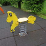 Na zdjęciu znajduje się żółty bujak sprężynowy w kształcie konika, umieszczony na placu zabaw.