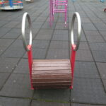 lement placu zabaw — niska beczka obrotowa w czerwonej ramie z uchwytami w formie metalowych okręgów po bokach.