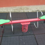 bujak sprężynowy w formie huśtawki wagowej z dwoma zielonymi siedziskami po bokach i czerwonym drążkiem umocowanym na dużej metalowej sprężynie.