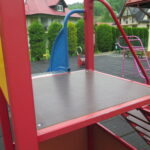 Platforma wieży zabaw o antypoślizgowej powierzchni, zabezpieczona kolorowymi panelami i barierkami ochronnymi, prowadzi do zjeżdżalni – całość wykonana z trwałych, bezpiecznych materiałów.