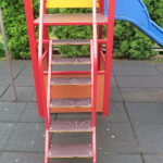 Widok z bliska na schodki prowadzące do wieży zabaw – metalowa konstrukcja, antypoślizgowe stopnie i kolorowe panele, wszystko ustawione na bezpiecznej nawierzchni z gumowych płyt.
