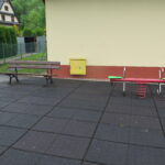 Fragment placu zabaw zlokalizowany przy budynku – widoczna równoważnia na sprężynie oraz ławka z oparciem, umożliwiająca odpoczynek.